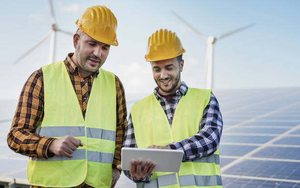 istock Men working with digital tablet at solar power station - Solar panels with wind turbines in background - Renewable energy concept 1320798079