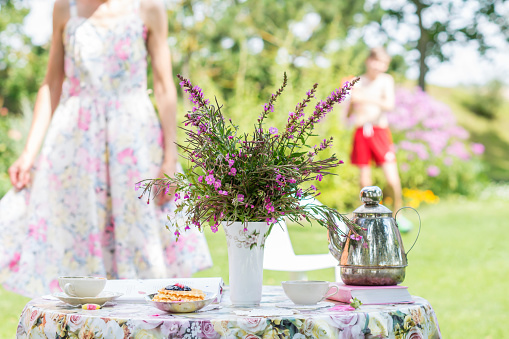 Wild flowers bouquet of  rosebay willowherbs or  fireweed in a vase on a flowery tablecloth , cake and teapot , cups with tea, woman and boy standing in background outdoors in summer time in the garden