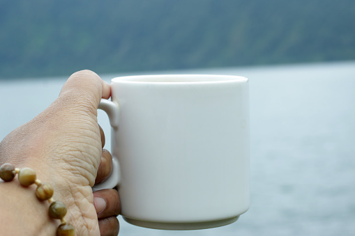 Person holding a blank white cup of coffee or tea in hand on blue nature lake background. Self love and care concepts. Business concept.