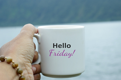 Friday coffee. Hello Friday greeting text on a white cup of coffee or tea on blue lake nature background. Weekend on vacation concept. Happy Friday.