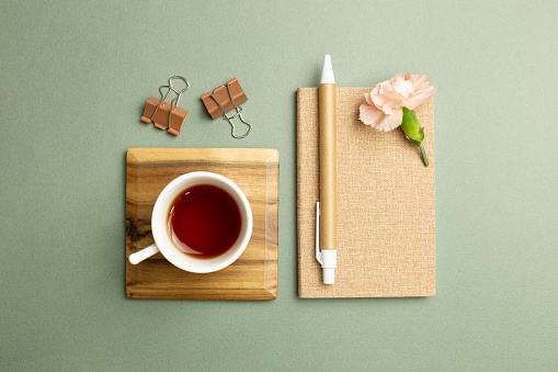 Notebook, cup of tea, clip, pen, carnation flower on green background. flat lay, top view, copy space. Work and study place