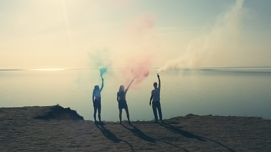 The three people with a color smoke sticks stand on the sea shore
