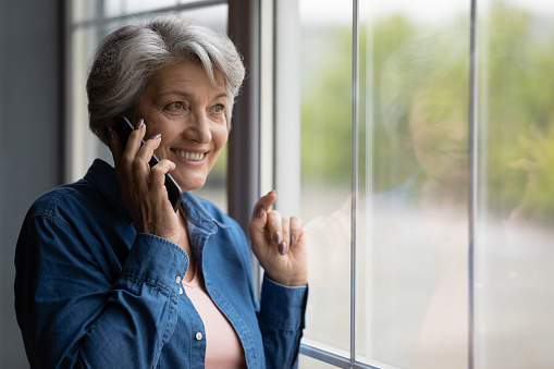 Mature 60s grey-haired woman standing at home looking out window talking on the mobile phone. Older generation and modern tech usage, phonecall activity, make order and communication distantly concept