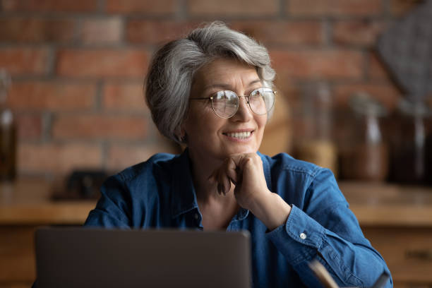 idosa senta na cozinha perto de laptop sorriso olhar para longe - pensive women business mature adult - fotografias e filmes do acervo