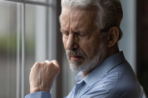 Close up sad older 70s man standing near window with eyes closed. Lonely vulnerable abandoned bearded grandfather suffer about disorder, incurable senile disease, widower portrait, loneliness concept