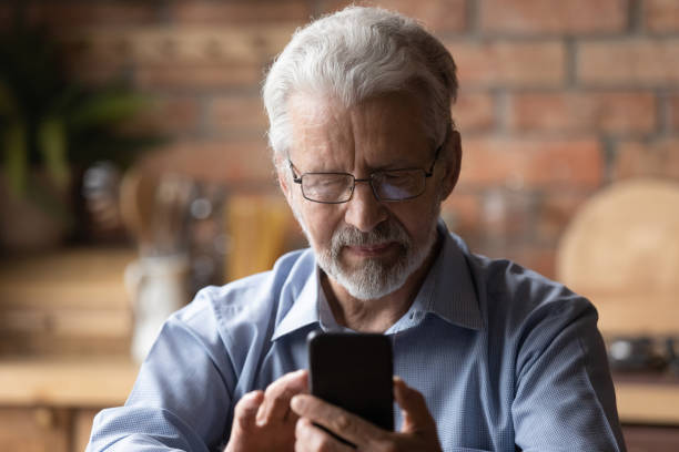 istock Serious mature man in glasses sit in kitchen holding smartphone 1320817191