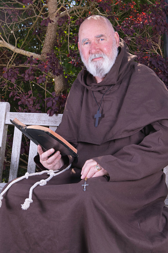 Older Franciscan monk reading his bible in a quiet spot in a garden.