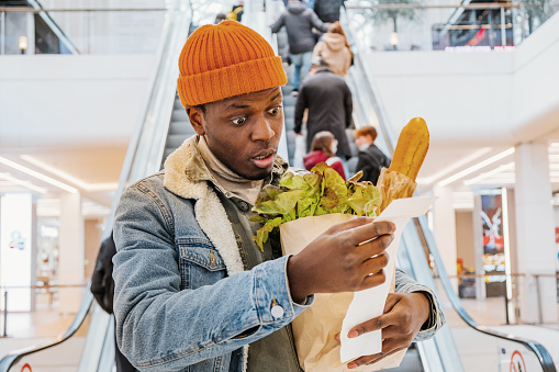 African man with a paper bag of groceries looks surprised and upset at a receipt from a supermarket with high prices against the background of an escalator with customers in the shopping center. The rise in the price of food.