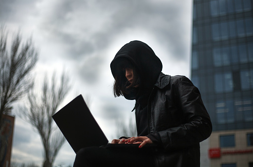 Girl in a black leather coat and a hood with laptop on a city street, concept of a woman hacker and agent working remotely at computer