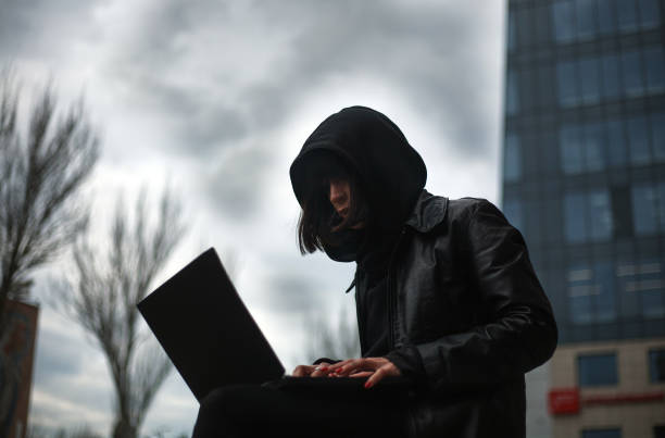 Girl in black leather coat and hood with laptop on city street, concept of woman hacker and agent working remotely at computer Girl in a black leather coat and a hood with laptop on a city street, concept of a woman hacker and agent working remotely at computer computer hacker spy computer crime laptop stock pictures, royalty-free photos & images