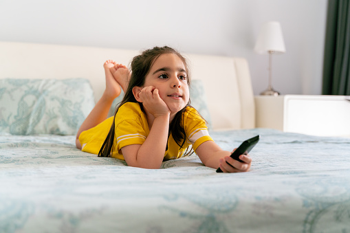 Little girl watching TV lying on bed with remote control in hand