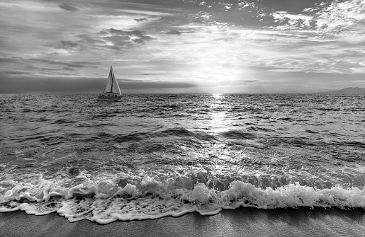 A Sailboat Is Sailing Out To Sea As a Gentle Wave Rolls to Shore In Black And White Image Format
