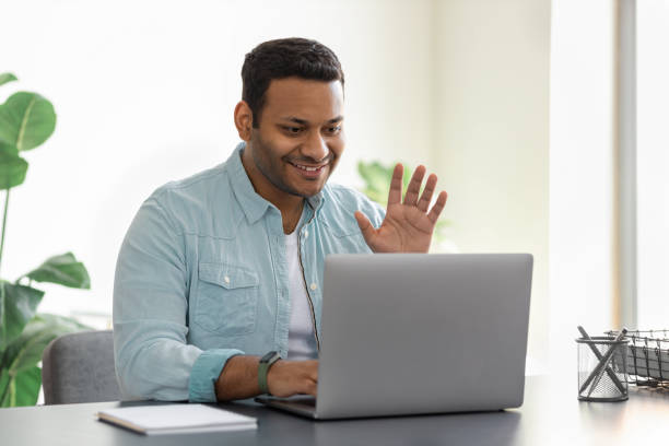 istock Friendly young Indian man using laptop for video communication with employees. Male freelancer in casual clothes waving hand in front of webcam greeting online participants 1320888838
