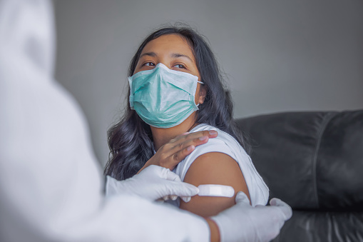 Close-up shot of unrecognizable Southeast Asian doctor sticking adhesive bandage to patient's arm. The doctor wearing a surgical glove for protection while the patient wearing a surgical mask.