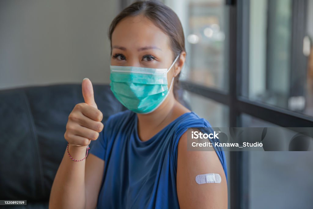 Proud Asian Woman Giving Thumbs Up After Receiving a COVID-19 Vaccine Close-up shot of proud Asian woman with protective face mask giving a thumbs up after receiving a COVID-19 vaccine at medical clinic. She's smiling behind her mask after a success vaccination. COVID-19 Vaccine Stock Photo
