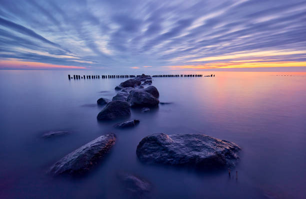 The IJsselmeer in the Netherlands during sunrise. In the water is a sort of path of big boulders leading into the water towards a row of small poles. The sky is filled with thin clouds and the sun shines brightly through them. The water is very peaceful and reflects the sky.
