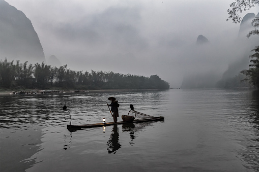 Chinese senior fisherman floating on his wooden raft with two gray herons on the Li River. Early morning fog at sunrise. Rural chinese scene with senior chinese fisherman in the early morning mist.  Xing Ping, Yangshuo County, Guangxi, Guilin, China.
