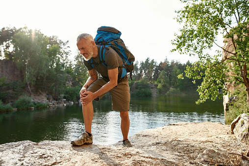 Tired middle aged caucasian man with dislocated knee hiking alone in summer nature. Hiking, travel outdoor, recreation and active adventure concept