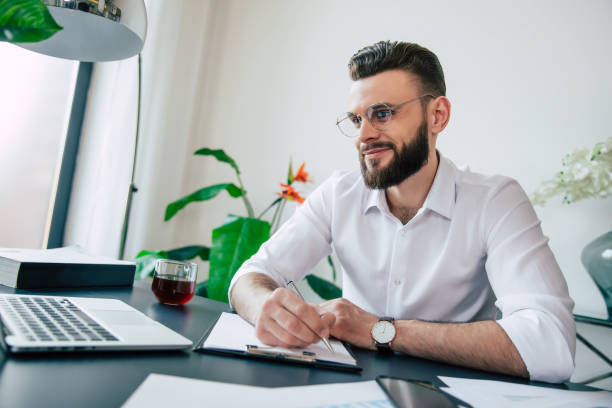 istock Young modern handsome stylish bearded businessman in glasses and a white shirt is working on the laptop with documents, papers on the desk of the office workplace. 1320899607