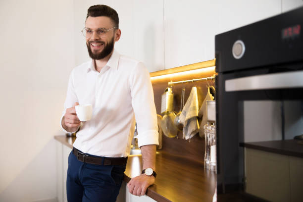 relaxed bearded confident businessman in smart casual wear drinks smells coffee and thoughtful at domestic kitchen - business styles foods and drinks drinking imagens e fotografias de stock