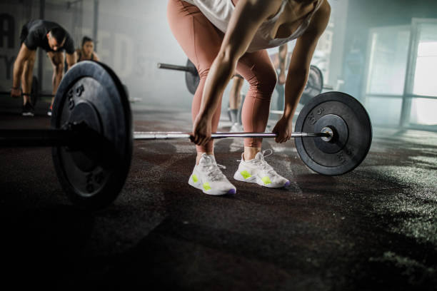 Unrecognizable female athlete exercising with barbell in a gym. Unrecognizable athletic woman having weight training with barbell in a gym. There are people in the background. Copy space. powerlifting stock pictures, royalty-free photos & images