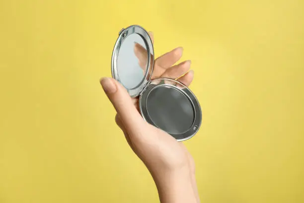 Woman holding metal cosmetic pocket mirror on yellow background, closeup