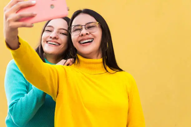 Photo of two pretty young smiling hispanic women in blue and yellow sweaters with braces on their teeth taking a selfie. friendship concept.