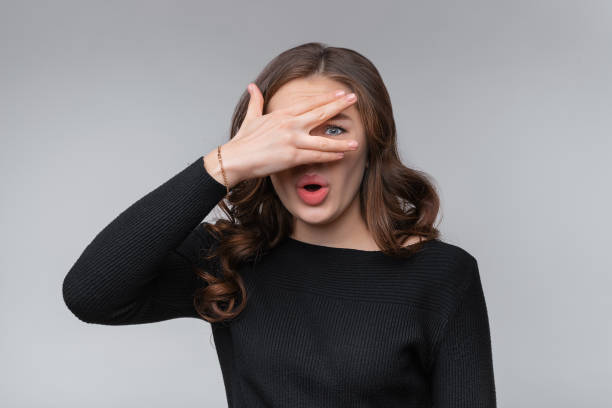 Portrait of scared and anxious young brunette woman, covering eyes with hand, peeking through fingers worried, panicking Portrait of scared and anxious young brunette woman, covering eyes with hand, peeking through fingers worried, panicking, standing on grey background hair covering eyes stock pictures, royalty-free photos & images