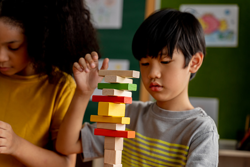 ocused Asian boy building tower with wooden blocks while playing near African American girl in school