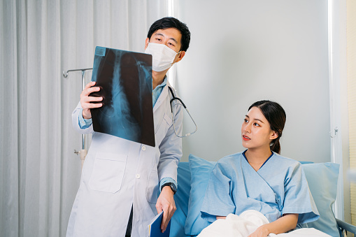 Young Asian male doctor with face mask and stethoscope in labcoat explaining and reviewing x-ray report to patient sitting in hospital bed