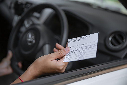 Close-up shot of unrecognizable Asian woman in a car holding a COVID-19 vaccination record card at drive-up clinic