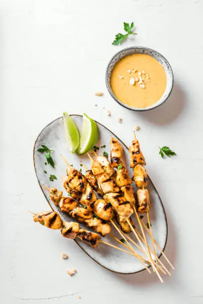 Chicken grilled satay skewers served with lime and peanut sauce on white, top view. Asian thai style food.