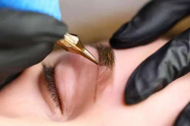 Close-up of professional makeup artist using golden tool and applying colour on eyebrow shape. Macro shot of specialists hands in protective gloves. Beauty and permanent procedure concept