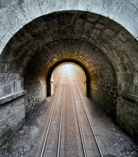 istock Dark passageway of roughly hewn stones with a round arch through which run two rail lines, composite surreal image 1320946466