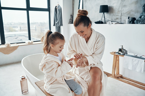 Mother with daughter in bathrobes smiling and using hand cream while doing morning routine