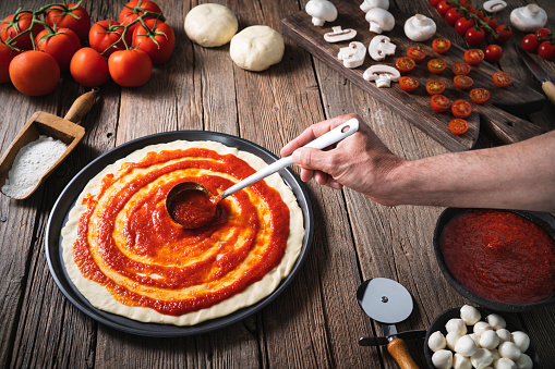 Italian pizza male hand spreading tomato sauce with ladle on the pizza raw dough with ingredients as mozzarella, mushrooms and tomato on rustic wooden table