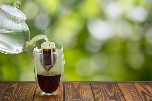 hand drip coffee. Hot water pouring from flying teapot into filter bag in glass on wooden table and green blurred background.