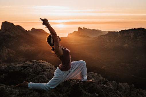 Young girl doing the yoga upward salute Urdhva Hastasana position what is part of the sun salute during the sunrise in a mountainous landscape. Color editing with slightly grain. Part of a series.