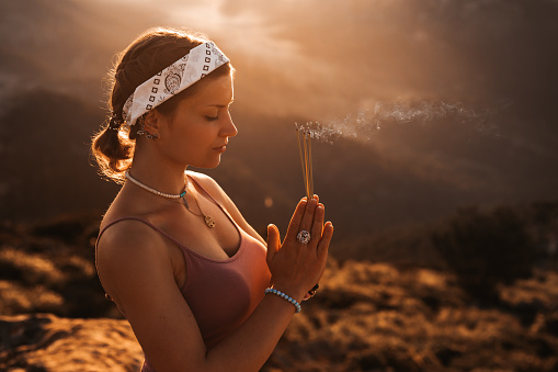 Beautiful girl doing meditation with incense sticks and closed eyes during the sunrise in a mountainous landscape in Majorca. Color editing with slightly grain. Part of a series.