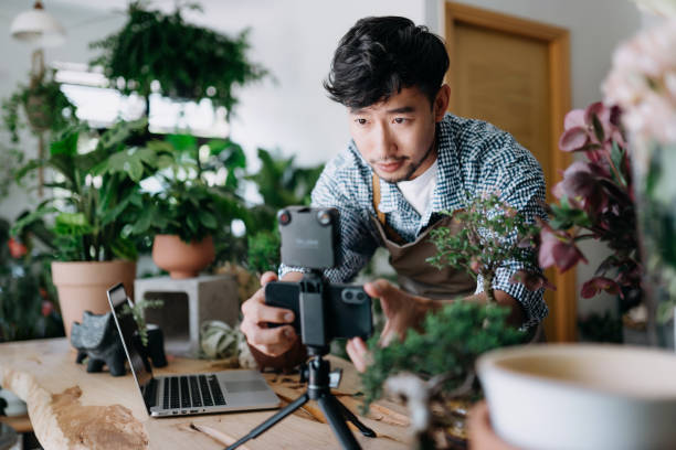 年輕的亞洲男性植物學家，小企業花店的老闆，在開始在線盆栽植物安排教程之前，先設置智能手機照明，在花店的智慧手機和筆記型電腦上寫博客。電子學習、在線教程概念 - choicesea 個照片及圖片檔