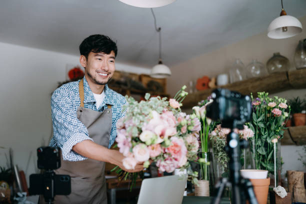 молодой азиатский ботаник-мужчина, владелец цветочного магазина малого бизнеса, vlogging на камеру и ноутбук, делая онлайн учебник по цветочны� - florist flower arranging male стоковые фото и изображения