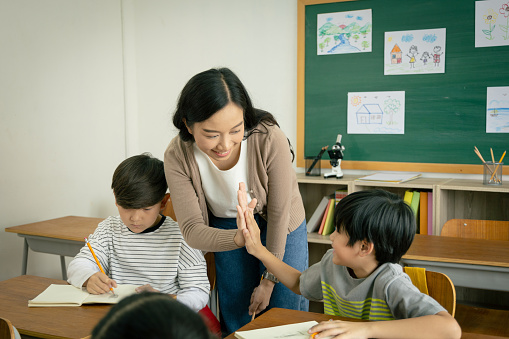 A young Asian female teacher and an elementary school boy clapping together hands in the classroom. The teacher congratulates the students on the success