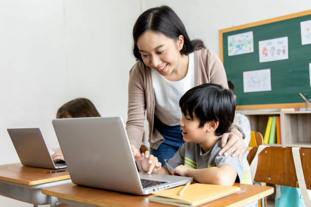 An Asian female teacher teaches primary school students to use computers and tablets to search for knowledge and information through the Internet. Classroom diversity students. Back to school concept stock photo
