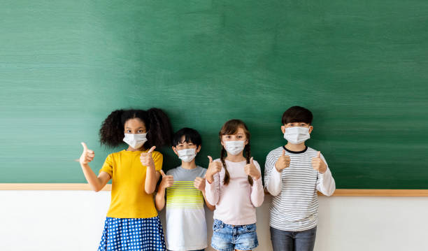 Diversity of children students wearing medical masks in the classroom. Students stand in a row in front of the blackboard with thumbs up.  Back to School And new normal Concept stock photo