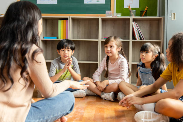 le studentesse delle scuole elementari diversity che siedono al piano della classe ascoltano le insegnanti asiatiche raccontare storie. concetto di istruzione e apprendimento - floor sitting foto e immagini stock