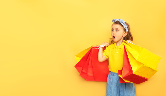 Surprised child girl in summer clothes hold package bag with purchases isolated on yellow background studio. Shopping discount sale concept. Mock up,copy space, banner. Black friday