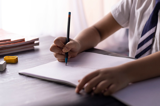 Selective focus on hand of an Indian child writing with pencil on notebook wearing school uniform