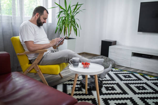 istock Robot cleaning floor while man sitting on sofa, using digital tablet at home and drink coffee 1320981956