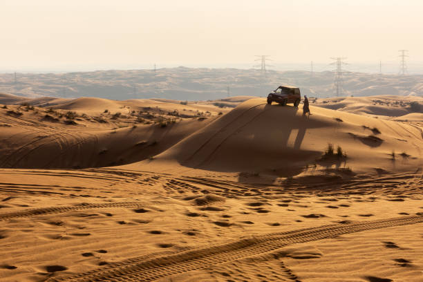 silhouette de voiture 4x4 et homme arabe en vêtements traditionnels au sommet d’une dune de sable au coucher du soleil, fossil rock, sharjah, émirats arabes unis. - 4x4 desert sports utility vehicle dubai photos et images de collection