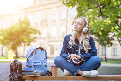 Positive happy cute student girl wearing denim jacket sitting on bench outdoors at campus, using mobile phone chatting. Blond young woman is listening music with headphones on university background.